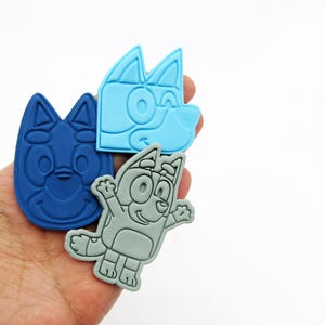 May include: Three cartoon character-shaped rubber charms in shades of blue and gray are held in a hand. The charms feature the faces and full body of a cartoon dog. The background is plain white.