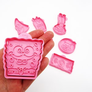 Könnte beinhalten: Rosa Kunststoff-Ausstecher in verschiedenen Formen, darunter SpongeBob Schwammkopf, Patrick Star und ein Geldschein. Die Ausstecher sind mit detaillierten Konturen versehen und eignen sich ideal zum Backen von thematischen Leckereien.