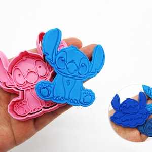 May include: Pink and blue Stitch-shaped cookie cutters. The blue cutter is held in a hand, with a pink cutter next to it. A smaller, circular image shows additional blue cutters with the text "Lilo & Stitch".