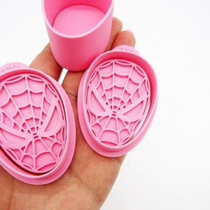 May include: Two pink plastic cookie cutters shaped like a Spiderman mask with a web design. The cutters are held in a hand.