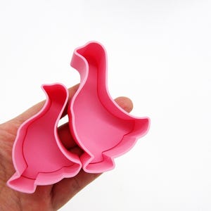Könnte beinhalten: Zwei rosa Ausstecher in Gänseform aus Kunststoff. Die Ausstecher werden in einer Hand vor weißem Hintergrund gehalten. Die Gänseformen sind detailliert mit einem gebogenen Hals und einem ausgestellten Boden, ideal zum Backen.