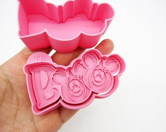 May include: Pink plastic cookie cutters shaped like the word "BOO" with two Mickey Mouse heads. The cutters are in the shape of a single piece, with the word "BOO" on the left and the Mickey Mouse heads on the right.