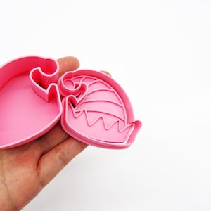 May include: Two pink plastic cookie cutters shaped like a Christmas elf hat. The cutters are held in a person's hand.