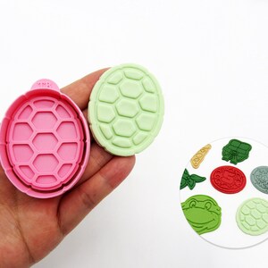 May include: A pink turtle-shaped cookie cutter and a light green turtle-shaped cookie. A small plate shows various green, red, and gray turtle-themed cookie designs. The cookie cutters and designs are for baking.