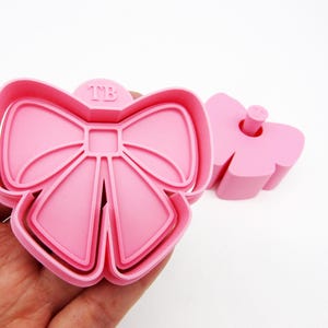May include: Pink plastic cookie cutter shaped like a bow with the letters "TB" embossed on the top. The cutter has a separate piece that fits inside to create a raised design.