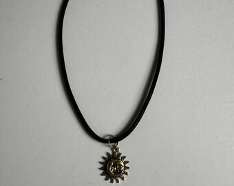May include: A black choker necklace with a gold-colored sun pendant. The sun pendant has a detailed face and rays. The necklace has a simple clasp.