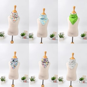 May include: A collection of six triangular baby bibs displayed on mannequins. The bibs feature various patterns including colorful fruits, light blue shapes, solid lime green, blue floral, gray geometric, and black and white abstract designs.