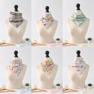 May include: A collection of six triangular baby bibs displayed on mannequin torsos. The bibs feature various patterns including grey deer, colorful floral, green geometric, multi-colored dinosaurs, jungle animals, and pastel unicorns on white fabric.