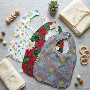 May include: Three baby bibs featuring distinct patterns: one with colorful cartoon monsters, one with red and white poinsettia flowers on a dark green background, and one with gray space-themed rockets and planets, accompanied by two folded muslin blankets and wooden rings.