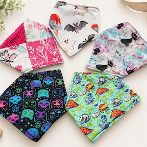 May include: Five triangular fabric bibs featuring diverse patterns: one with pink floral and dragonfly motifs, one with white ghosts and bats, one with black cats on gray, one with neon cat faces on black, and one with colorful dinosaurs on light blue.