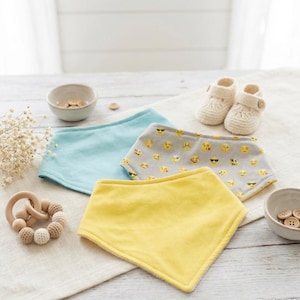 May include: A collection of baby items, including three bibs in blue, yellow, and gray with an emoji pattern. Also shown are a pair of cream-colored crochet baby shoes, a wooden teether, and small bowls with wooden buttons.