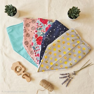 Könnte beinhalten: Fünf Baby-Lätzchen mit verschiedenen Mustern: hellblau, rosa mit Kreismuster, rot floral, marineblau mit Sternen und Planeten sowie grau mit gelben Smileys. Ein Holzrassel und kleine Sukkulenten sind ebenfalls vorhanden.