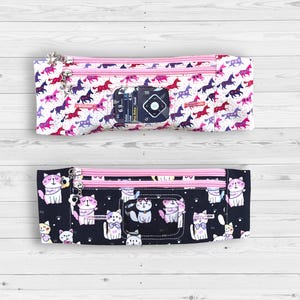 Puede incluir: Dos estuches rectangulares con cremallera. El estuche superior tiene un fondo blanco con patrones de unicornios morados, rosas y rojos. El estuche inferior tiene un fondo negro con patrones de gatos. Ambos estuches tienen cremalleras rosas.