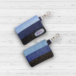 May include: Two denim keychains with silver-tone clasps. Each keychain features horizontal stripes of light blue, medium blue, dark blue, and black denim. One keychain has the name "ALVIJA" embroidered on a patch.