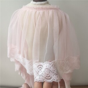 May include: A pink tulle cape with a white lace trim. The cape is gathered at the neck and has a flowing, ethereal look.