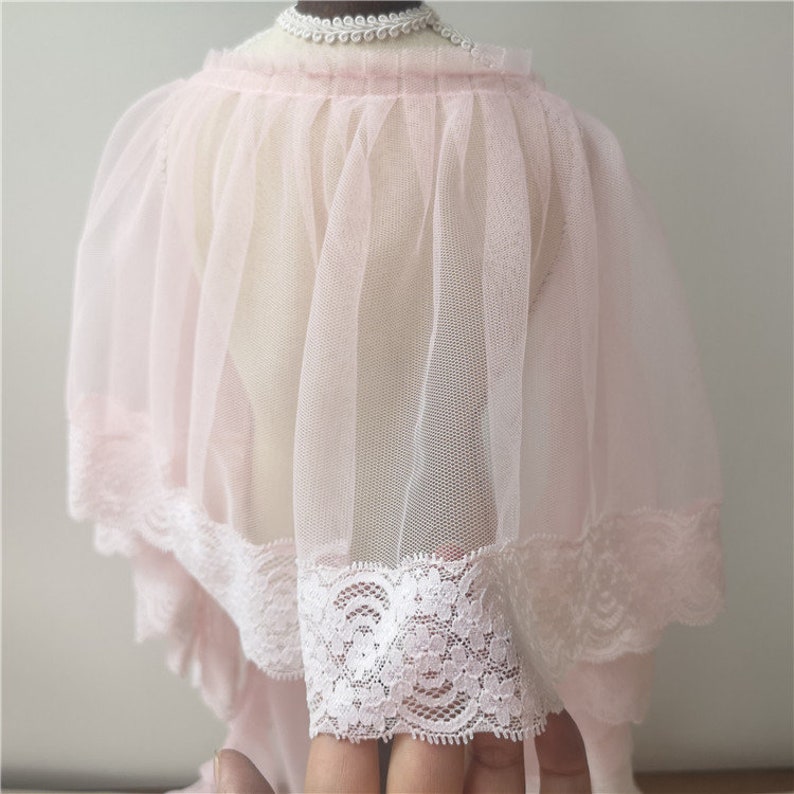 May include: A pink tulle cape with a white lace trim. The cape is gathered at the neck and has a flowing, ethereal look.