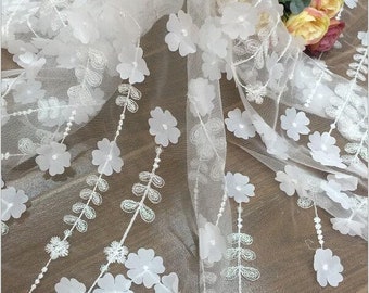May include: White floral lace fabric with 3D flowers and leaves. The fabric is sheer and delicate, with a soft, flowing texture.
