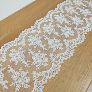 May include: White lace table runner with a delicate floral pattern. The runner has a scalloped edge and is made of a sheer fabric.