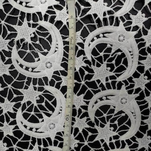 May include: White lace fabric with a repeating pattern of crescent moons and stars. The delicate design is set against a black background. A measuring tape shows the fabric's dimensions.