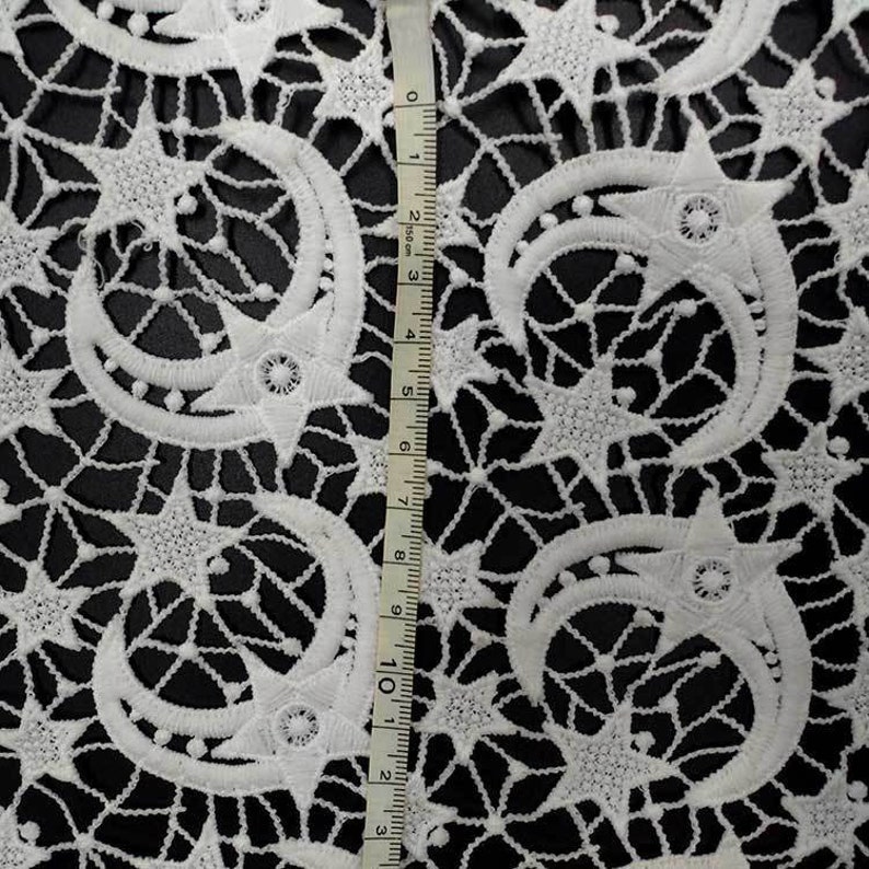 May include: White lace fabric with a repeating pattern of crescent moons and stars. The delicate design is set against a black background. A measuring tape shows the fabric's dimensions.