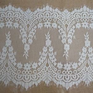 K&ouml;nnte beinhalten: Wei&szlig;e Spitzenborte mit einem gewellten Rand und einem Blumenmuster. Die Spitze ist zart und filigran, mit einer weichen, flie&szlig;enden Textur.