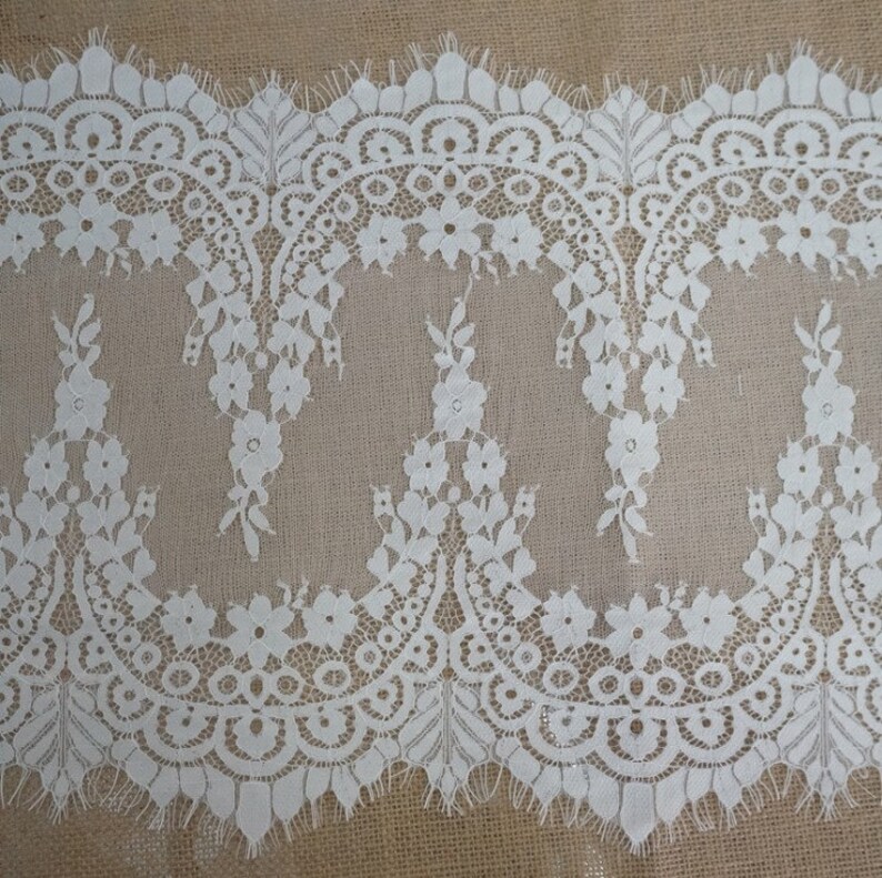 K&ouml;nnte beinhalten: Wei&szlig;e Spitzenborte mit einem gewellten Rand und einem Blumenmuster. Die Spitze ist zart und filigran, mit einer weichen, flie&szlig;enden Textur.
