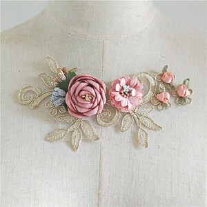 May include: A gold lace applique with pink and blue fabric flowers. The applique features a large pink rose, a smaller pink flower with a center of blue and white beads, and three smaller pink roses.