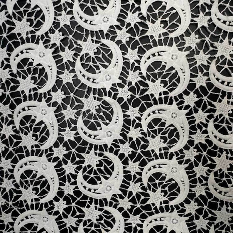 May include: White lace fabric with a repeating pattern of crescent moons and stars. The delicate design features intricate details and a contrasting black background, creating a celestial theme. This textile is suitable for various craft projects.