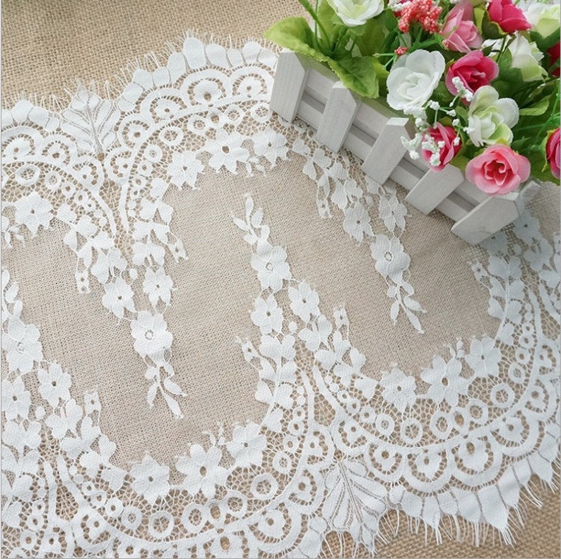 K&ouml;nnte beinhalten: Wei&szlig;e Spitzenborte mit einem gewellten Rand und einem floralen Muster. Die Spitze ist zart und filigran, mit einer weichen, flie&szlig;enden Textur.