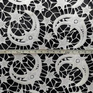 May include: Close-up of white lace fabric with a celestial design. The pattern features crescent moons and stars, with a ruler showing measurements in inches and centimeters. The lace is set against a black background.