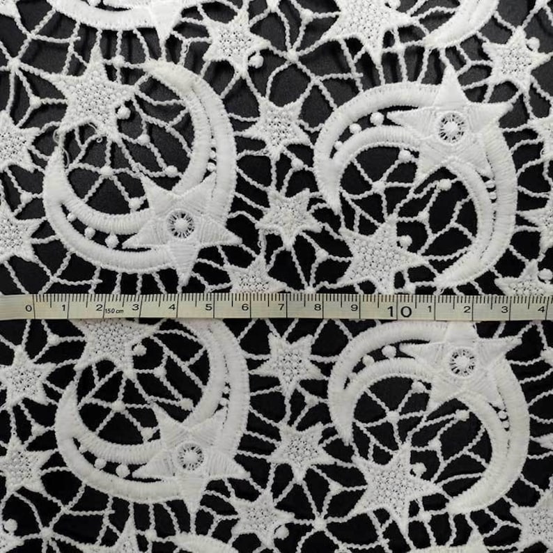 May include: Close-up of white lace fabric with a celestial design. The pattern features crescent moons and stars, with a ruler showing measurements in inches and centimeters. The lace is set against a black background.