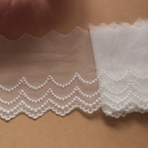 May include: Close-up of white lace trim with a scalloped edge. The lace features a delicate, dotted pattern along the wavy border. The trim is made of a sheer, net-like material, perfect for sewing and craft projects.