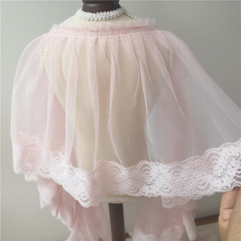 May include: A delicate pink tulle cape with a lace trim. The cape is sheer and has a flowing silhouette.