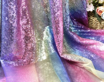 May include: A vibrant fabric with a rainbow gradient of colors, including pink, purple, blue, and yellow. The material is covered in small, reflective sequins, creating a shimmering effect. The fabric is draped, showing its texture and flow.