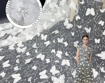 May include: White tulle fabric with a delicate butterfly pattern. The fabric is adorned with 3D butterfly appliques and small sequins. The fabric is shown draped over a table with a model wearing a dress with a similar pattern.