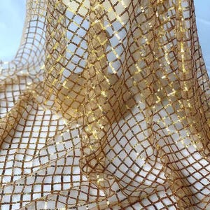 Könnte beinhalten: Nahaufnahme eines schimmernden, goldenen Stoffes mit einem Gittermuster aus Pailletten. Der Stoff ist drapiert und erzeugt Falten und Schatten, die die Textur und das Funkeln hervorheben. Das Material scheint leicht und für dekorative Zwecke geeignet zu sein.