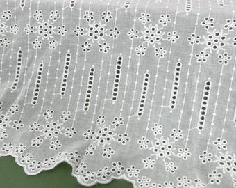 May include: White eyelet fabric with a scalloped edge. The fabric features a repeating pattern of floral and geometric cutouts and embroidered details. The fabric is likely cotton or a cotton blend, and is suitable for apparel or home decor.
