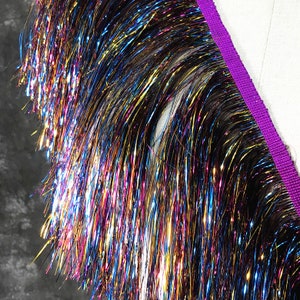 May include: A close-up of iridescent fringe trim with a purple backing. The fringe is made of thin, metallic strands that shimmer in the light.