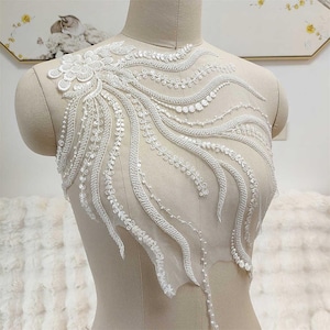 Könnte beinhalten: Weiße Perlen-Spitzenapplikation mit Blumenmuster. Die Spitze ist zart und filigran, mit einem fließenden Muster. Sie ist perfekt, um jedem Kleidungsstück einen Hauch von Eleganz zu verleihen.