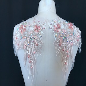 Może przedstawiać: Dwa delikatne kwiatowe aplikacje z opalizującymi kryształkami i różowymi perełkami. Aplikacje mają kaskadowy wzór z wiszącymi perełkami i koralikami.