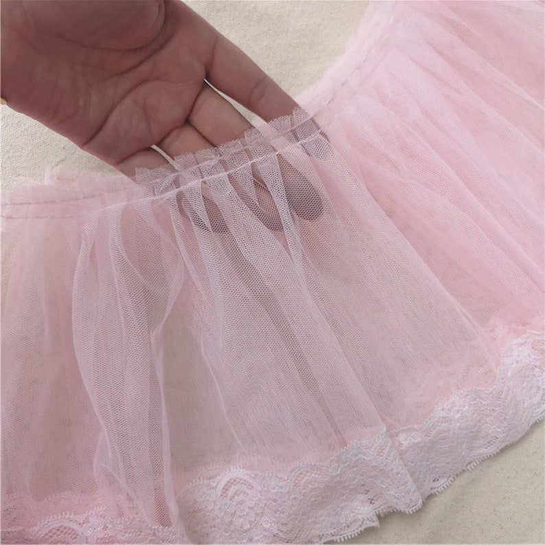 May include: Pink tulle fabric with a white lace trim. The tulle is gathered and ruffled, creating a delicate and feminine look.