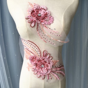 Może przedstawiać: Dwa różowe aplikacje kwiatowe z cekinami i kryształkami na białym tle. Aplikacje wykonane są z delikatnej tkaniny i charakteryzują się skomplikowanymi wzorami.