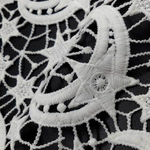 May include: Close-up of white lace fabric with a detailed, openwork design. The pattern features star and crescent moon shapes, creating a celestial theme. The lace is set against a dark background, highlighting the intricate details.