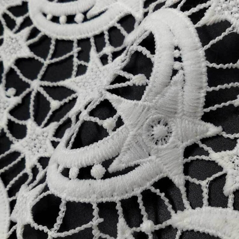 May include: Close-up of white lace fabric with a detailed, openwork design. The pattern features star and crescent moon shapes, creating a celestial theme. The lace is set against a dark background, highlighting the intricate details.