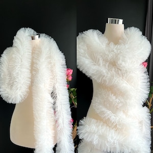 May include: White tulle fabric with a ruffled texture. The fabric is draped over a mannequin form, showcasing its delicate and airy quality.