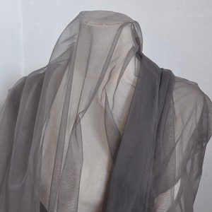 以下が含まれることがあります： マネキンにドレープされた、シアーグレーの生地。おそらくチュールまたはオーガンジーで、半透明で下の形が部分的に見えます。柔らかく流れるような外観の素材です。