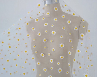 May include: Sheer white fabric with a daisy pattern. The fabric is draped over a mannequin, showcasing the delicate floral print. The daisies have white petals and yellow centers, creating a cheerful and airy design.