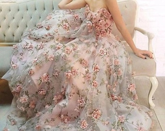 Op de afbeelding: Een strapless, lange jurk met een tule rok en 3D bloemenversieringen. De jurk is lichtroze met een lichtgrijze onderlaag.
