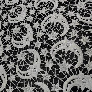 May include: White lace fabric with a repeating pattern of crescent moons and stars. The delicate design features openwork details, creating a textured appearance against a black background. The celestial theme is prominent in this decorative textile.