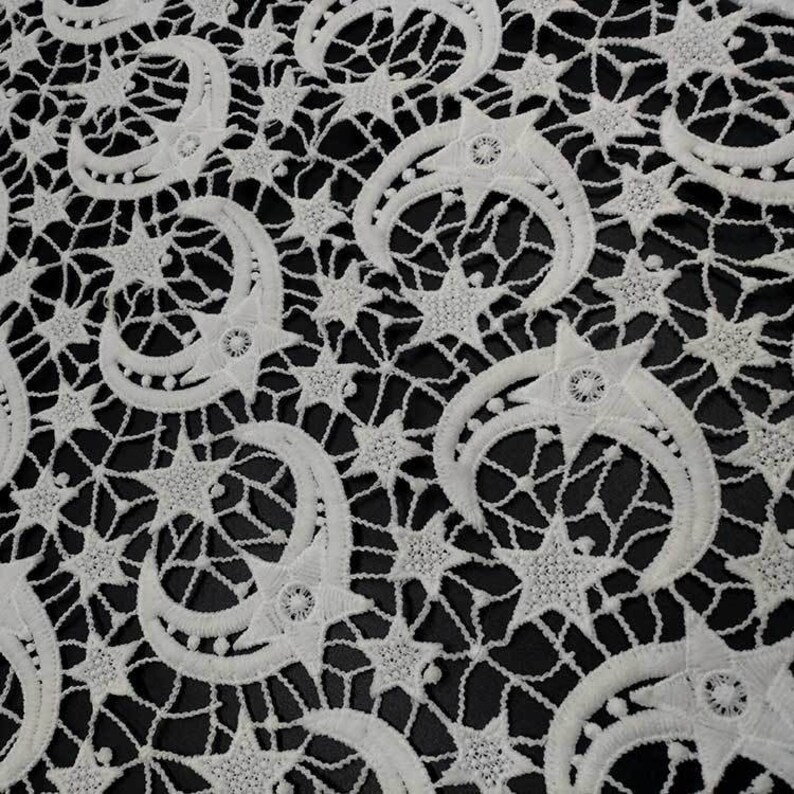 May include: White lace fabric with a repeating pattern of crescent moons and stars. The delicate design features openwork details, creating a textured appearance against a black background. The celestial theme is prominent in this decorative textile.